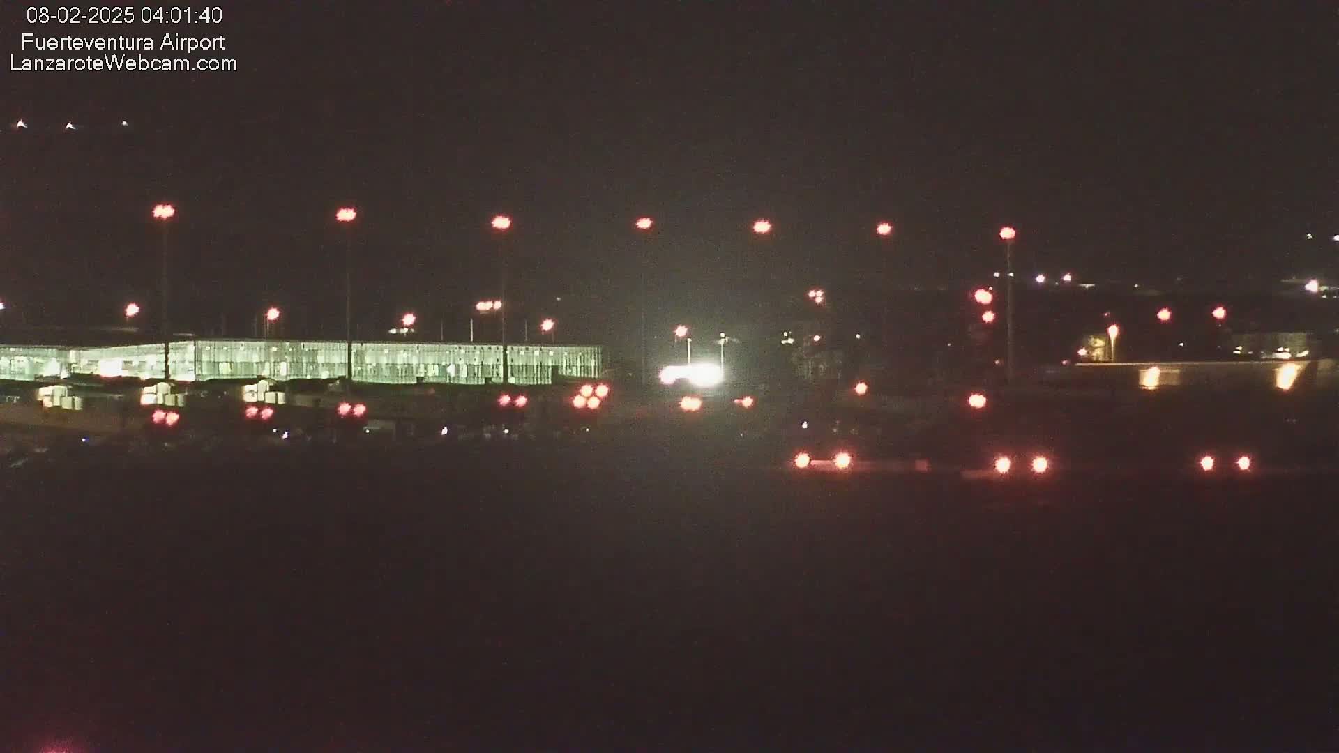 A nighttime view of an airport terminal building illuminated, with numerous red taillights visible in the foreground and several lights illuminating the tarmac.