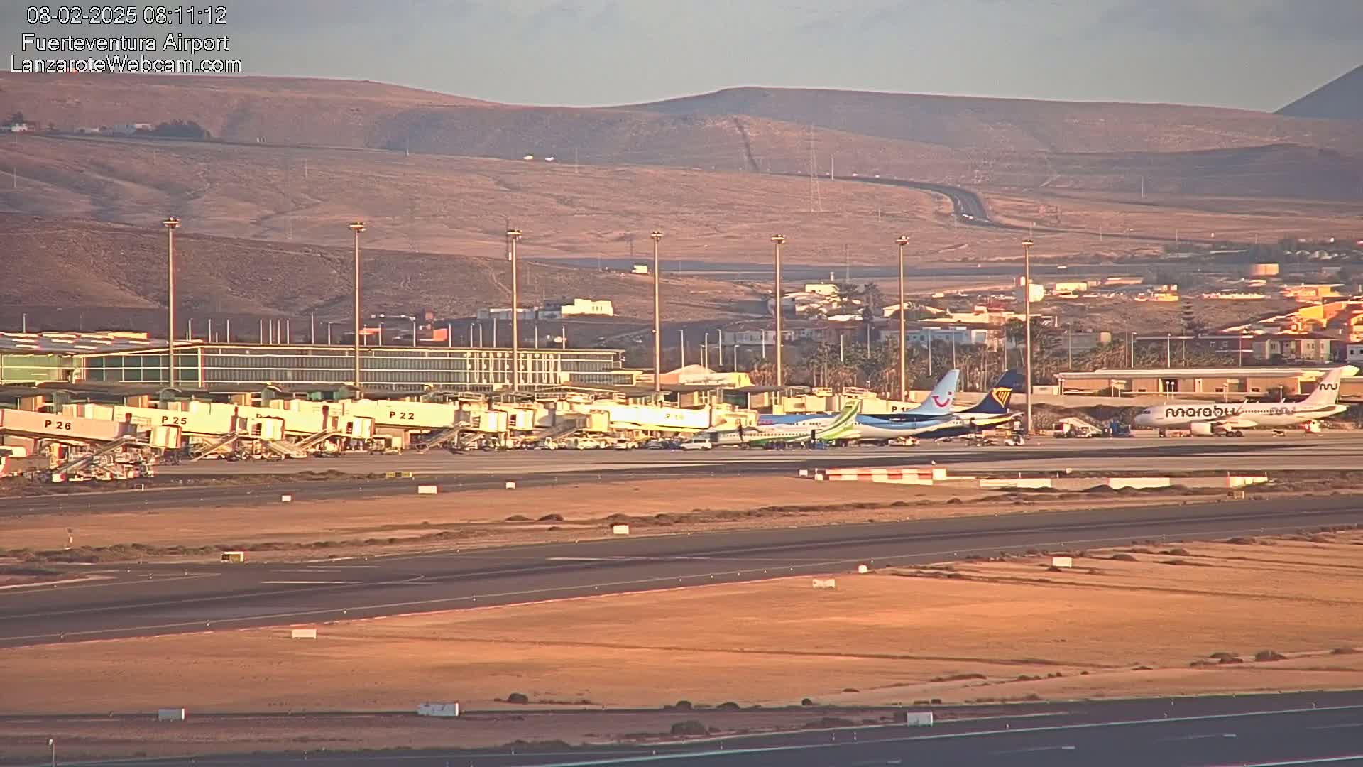 Several airplanes are parked at gates at an airport under a clear, light-orange sky, with a tan, hilly landscape in the background.