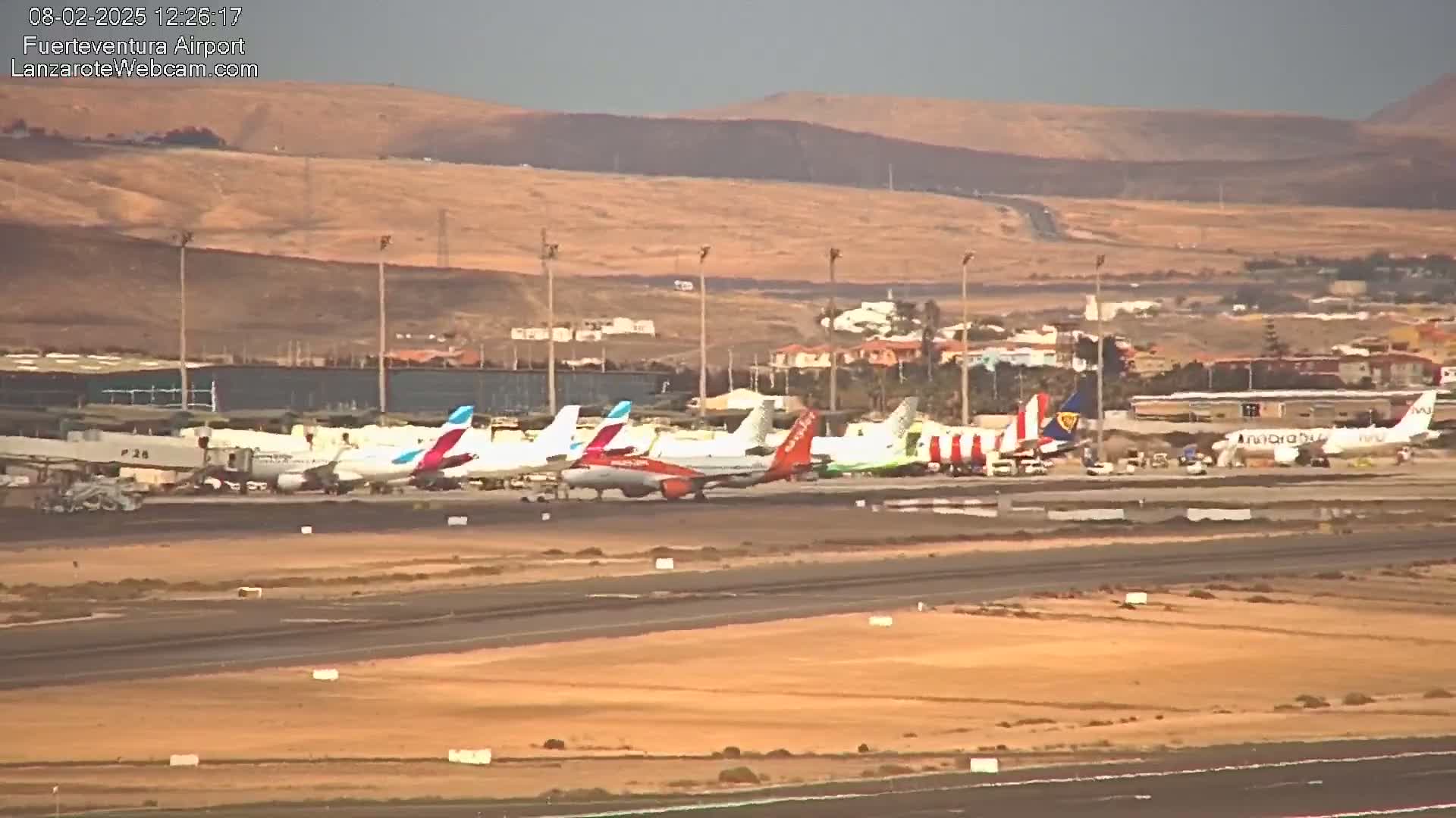 Several airplanes are parked at an airport tarmac under an overcast sky, with arid hills visible in the background.