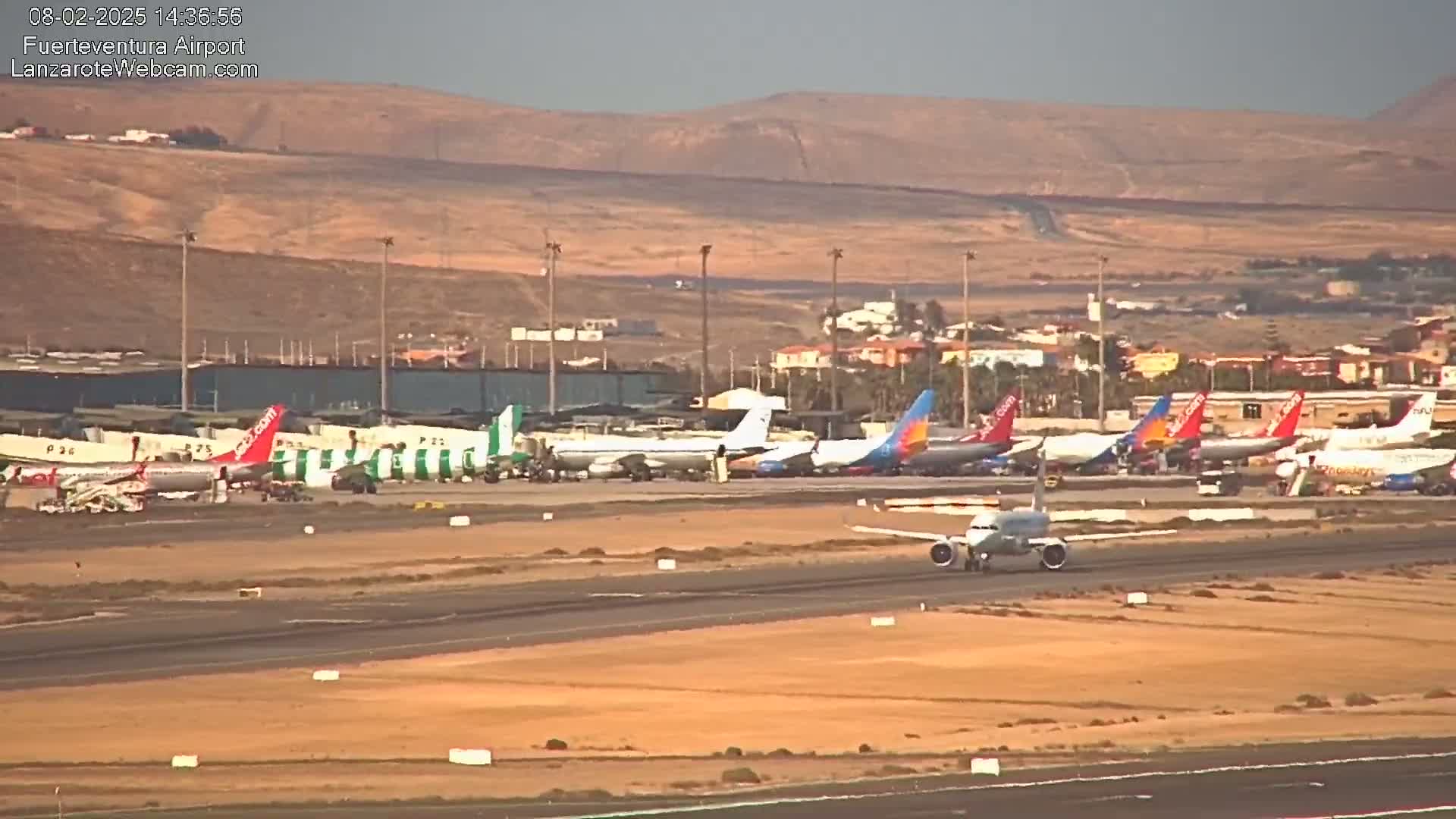 A plane taxis on a runway past numerous parked airplanes at an airport under a hazy, sunny sky.