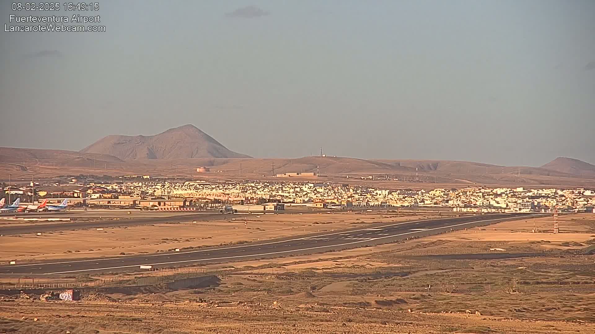 An airport runway stretches across a flat, arid landscape under a clear, light-blue sky, with a town and distant volcanic hills in the background.