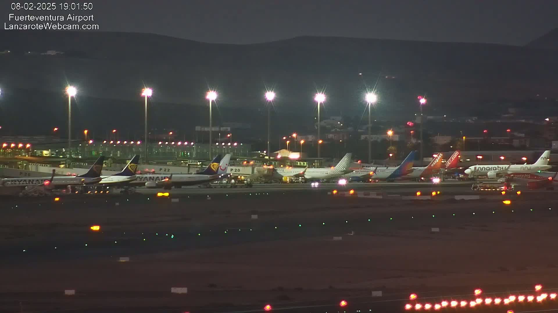 Several airplanes are parked on a tarmac at night under bright lights, with hills visible in the background.