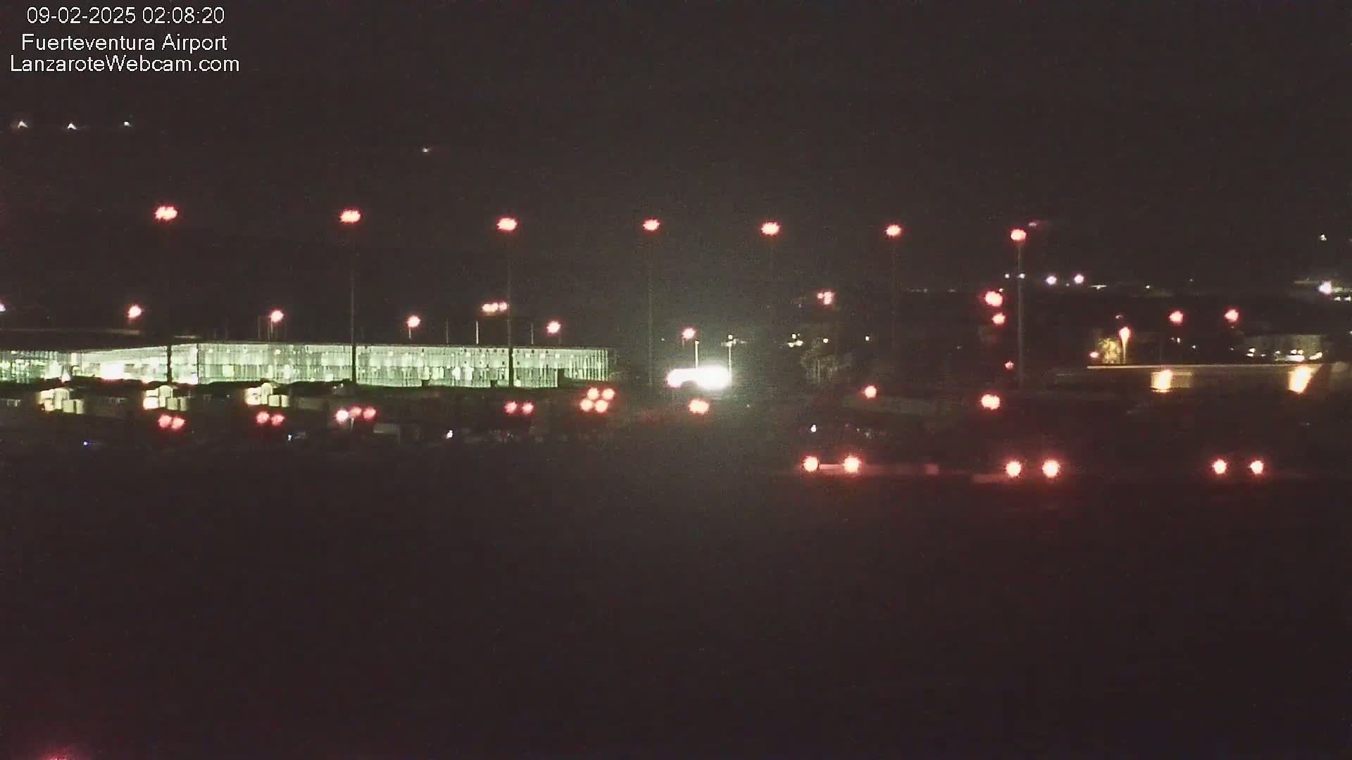 At night, a long, brightly lit building is visible, with vehicles and lights scattered in the foreground and background.