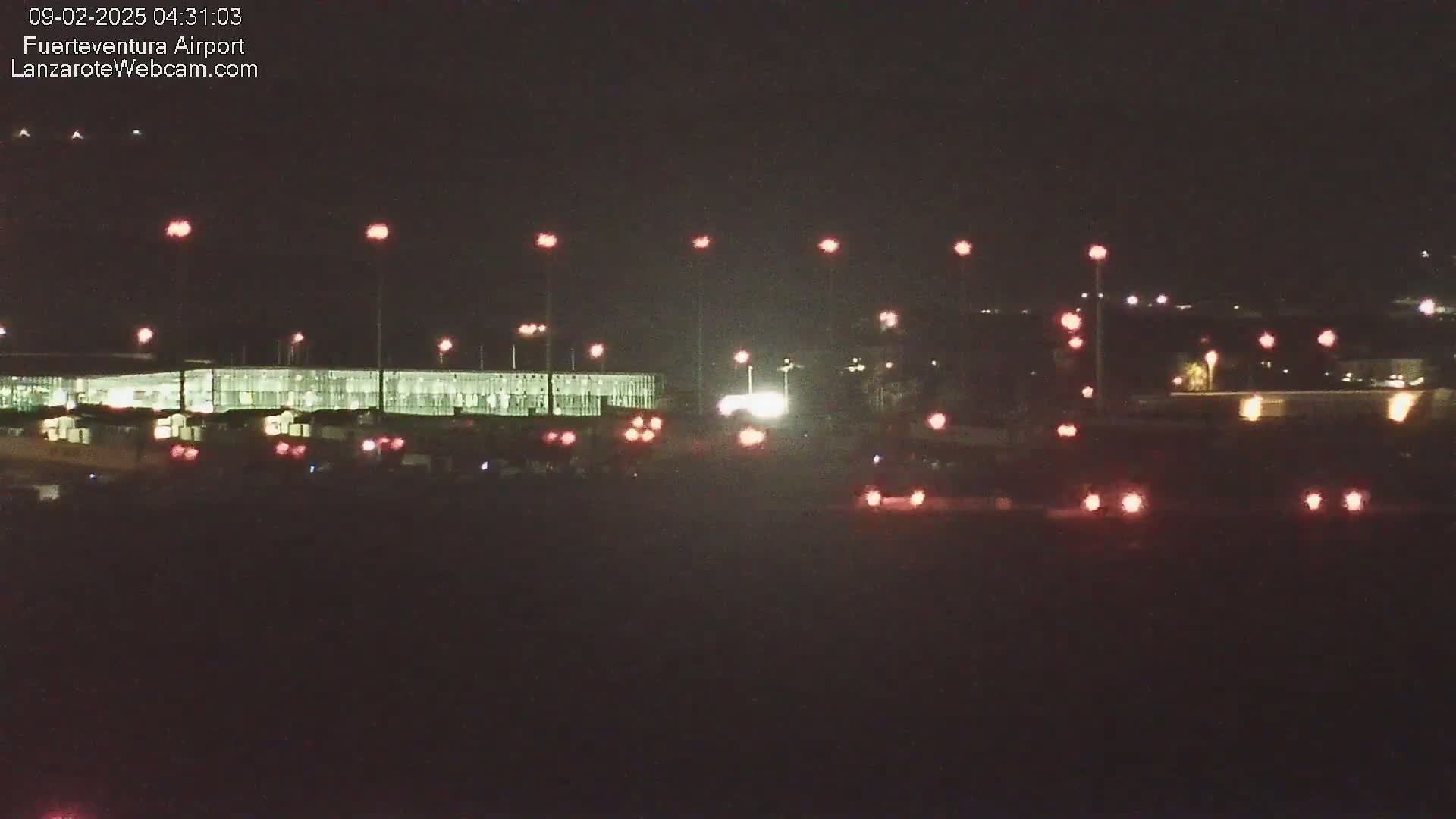 At night, a brightly lit airport terminal and roadway are visible, with numerous vehicles' taillights and roadway lights illuminating the scene.