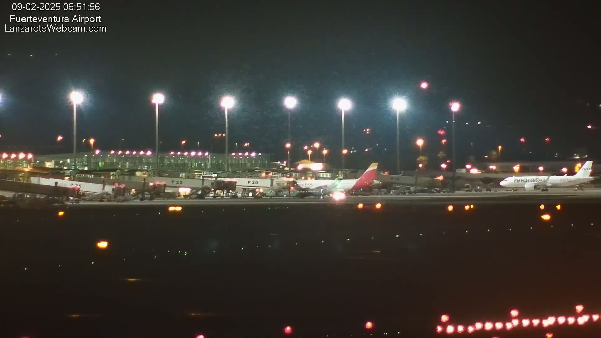 Several airplanes are parked at a brightly lit airport at night in light snow or rain.