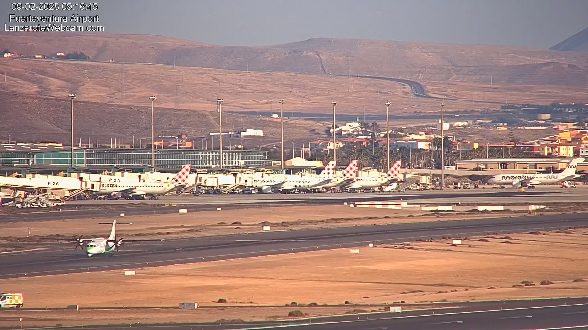 Several airplanes are parked at an airport tarmac under a clear, sunny sky with tan-colored hills visible in the distance.
