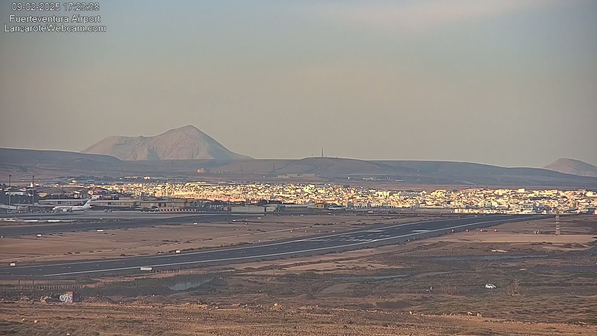 A long runway at an airport sits before a town and volcanic hills under a hazy sky.