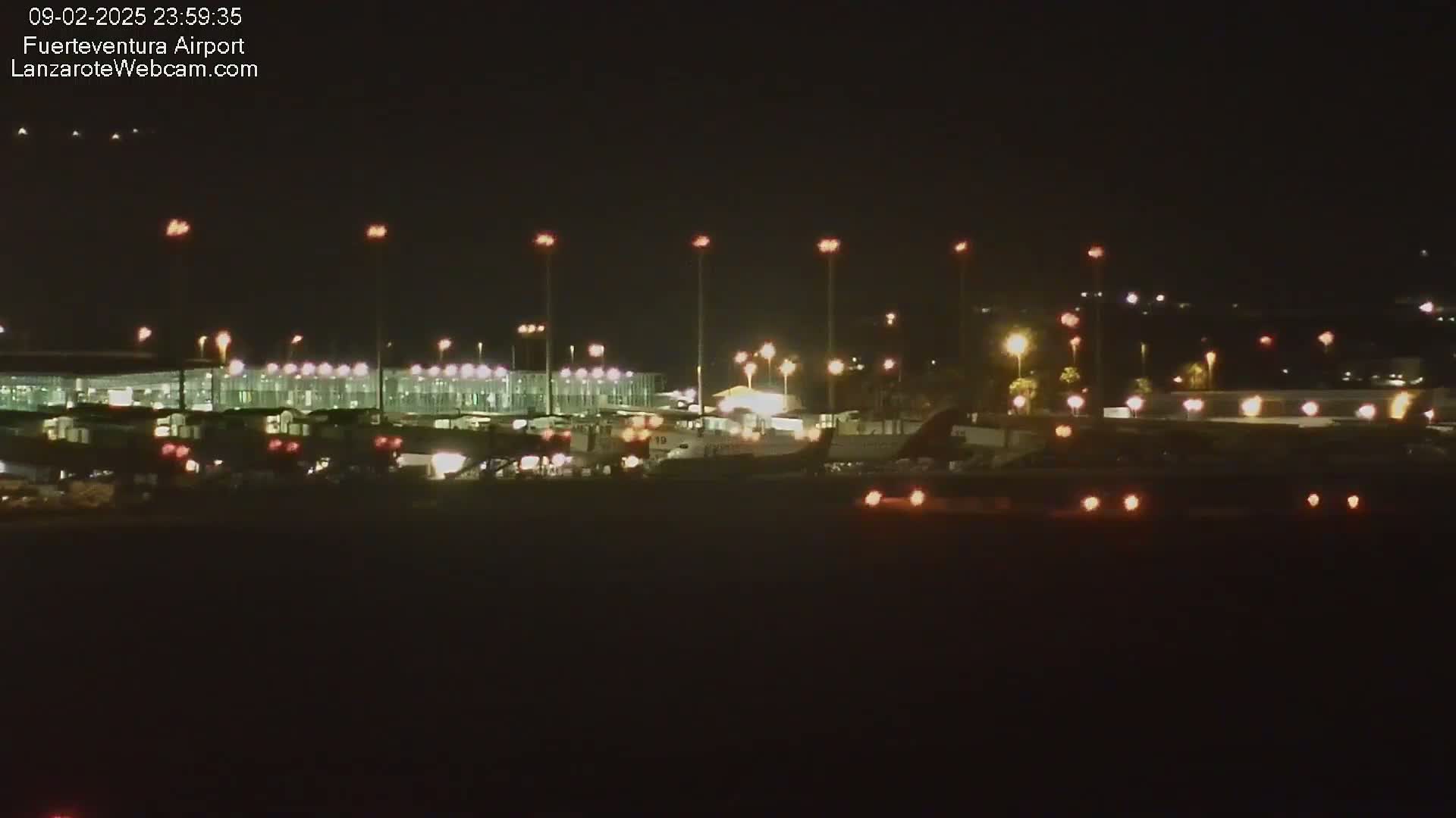 A nighttime long shot of an airport terminal and tarmac with airplanes and various ground vehicles visible under a clear, dark sky.