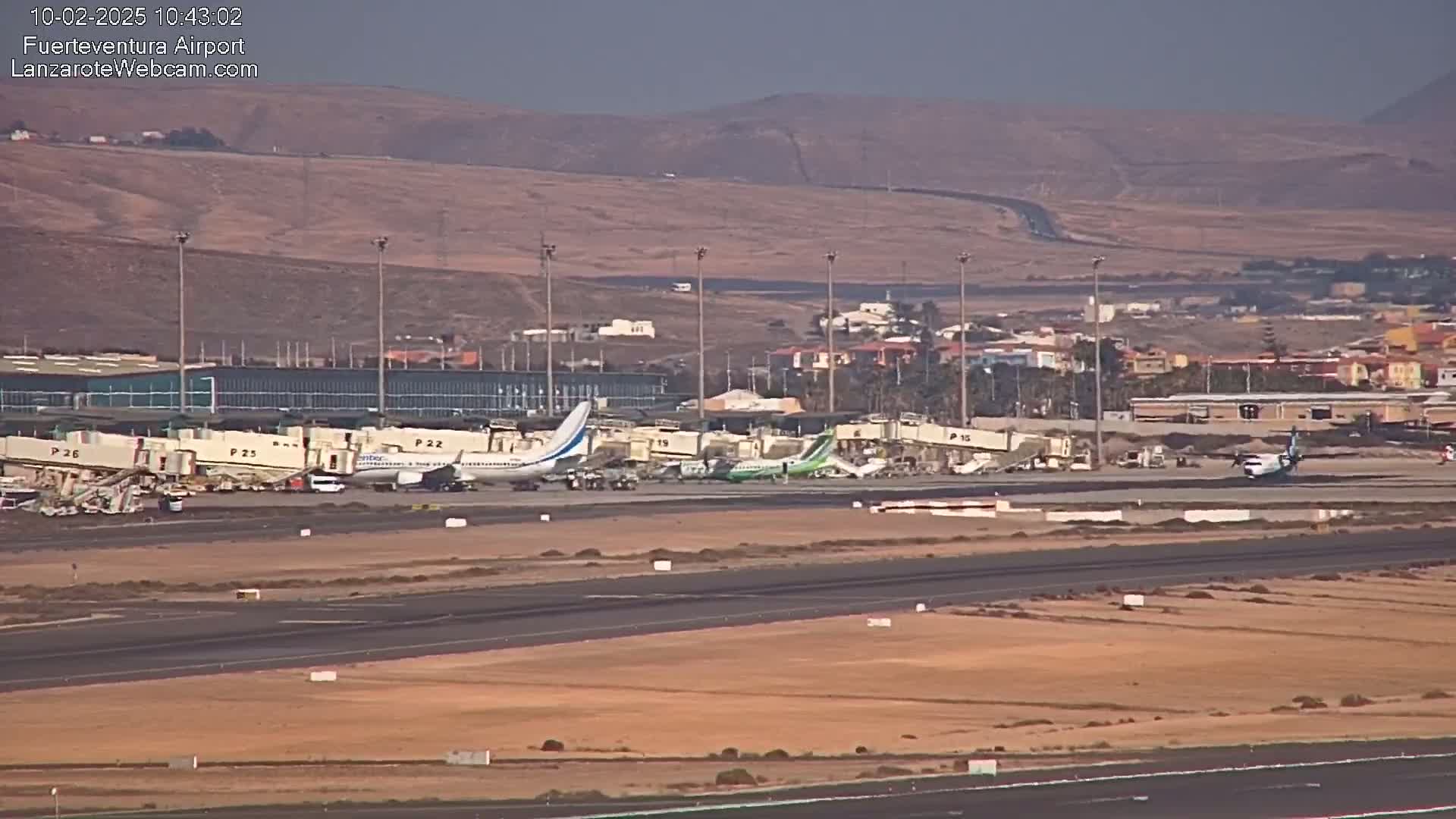 Several airplanes are parked at gates at an airport under hazy sunny conditions, with a town and hills visible in the background.