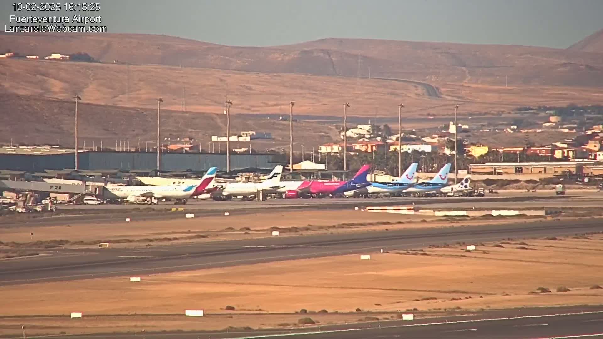 Several airplanes are parked on a tarmac at an airport under a hazy, sunny sky, with arid hills visible in the background.