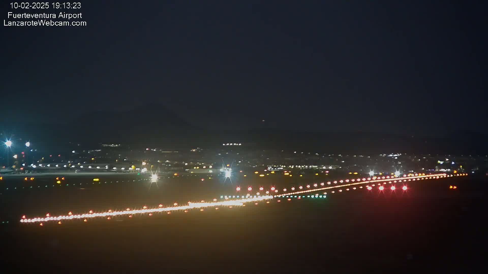 A nighttime long shot of a runway illuminated with lights, surrounded by dimly lit buildings and a dark sky.