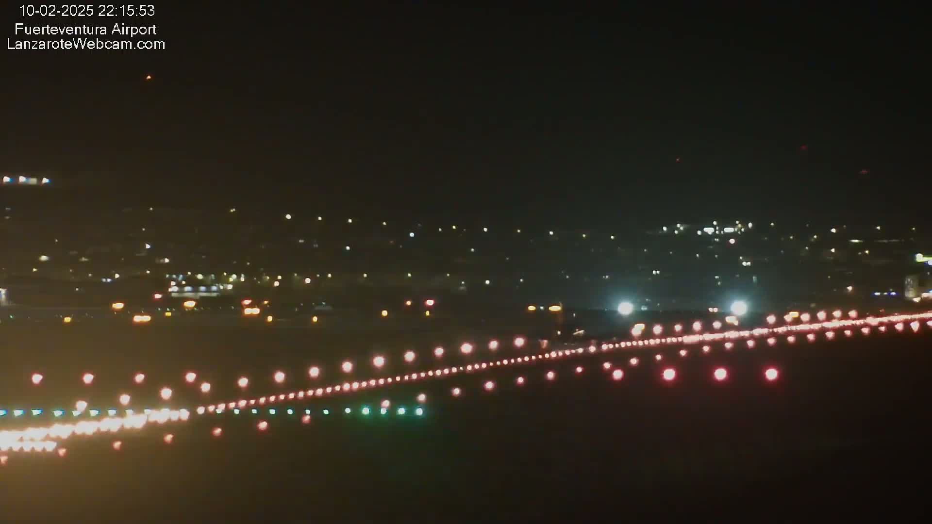 A nighttime long shot of an airport runway with lights illuminating the tarmac, a city skyline in the background, and a clear night sky.