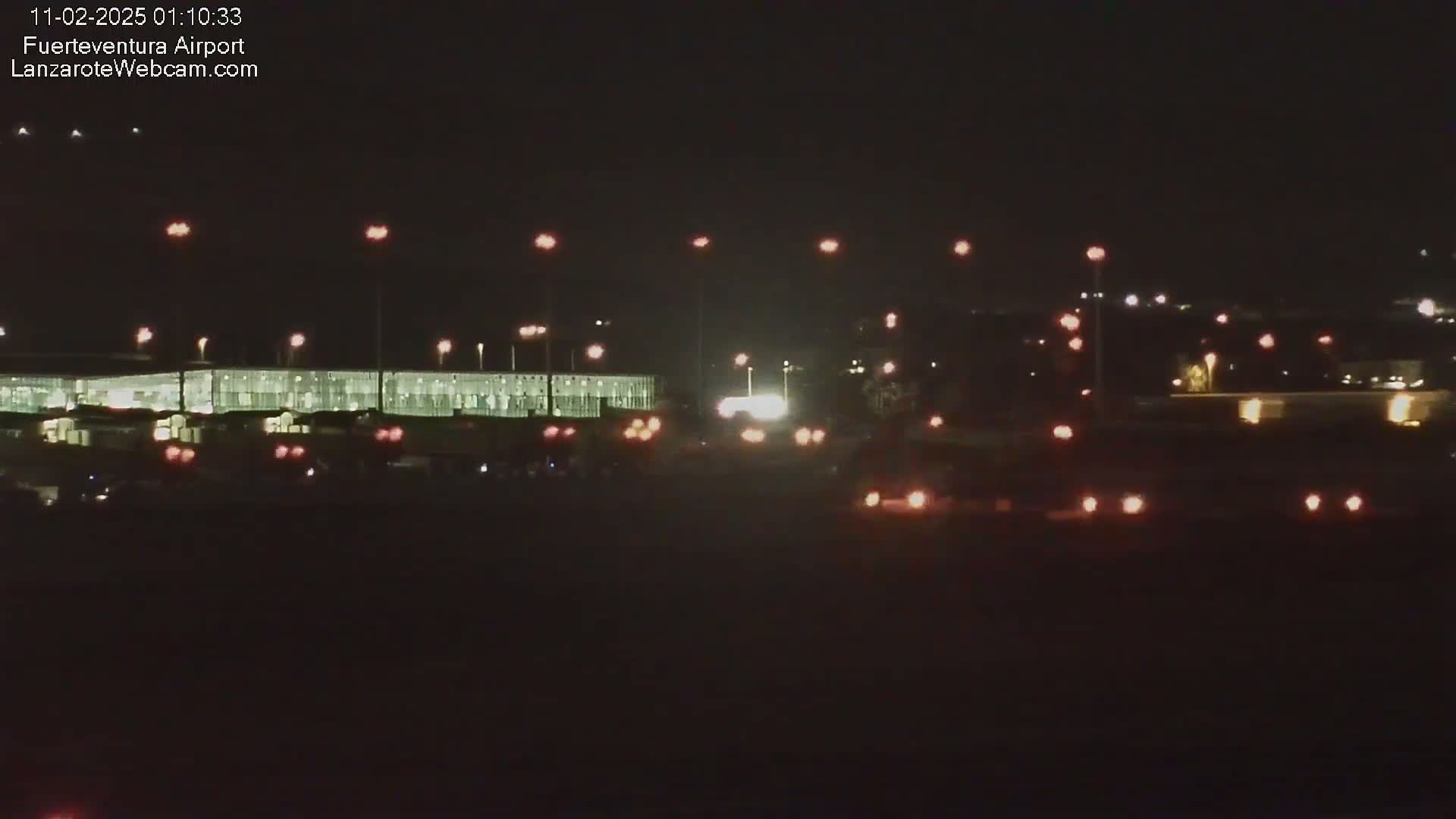 At night, a brightly lit airport terminal is visible with vehicles and lights scattered in the foreground and background.