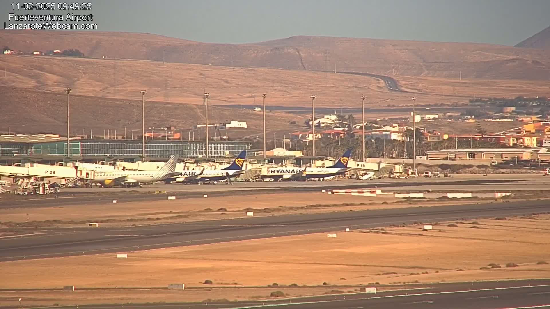 Several airplanes are parked at an airport tarmac under a clear, sunny sky, with a hilly, desert-like landscape visible in the background.
