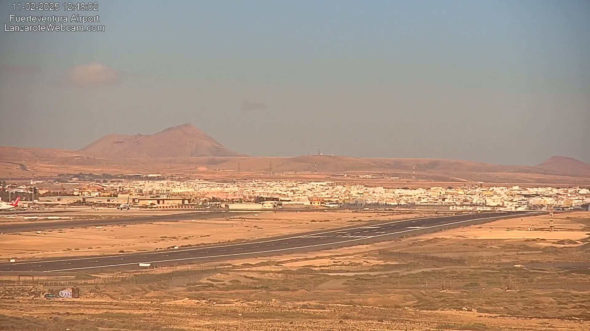 An airport runway and surrounding buildings are visible, with hazy or dusty atmospheric conditions and mountains in the background under a clear, light-colored sky.