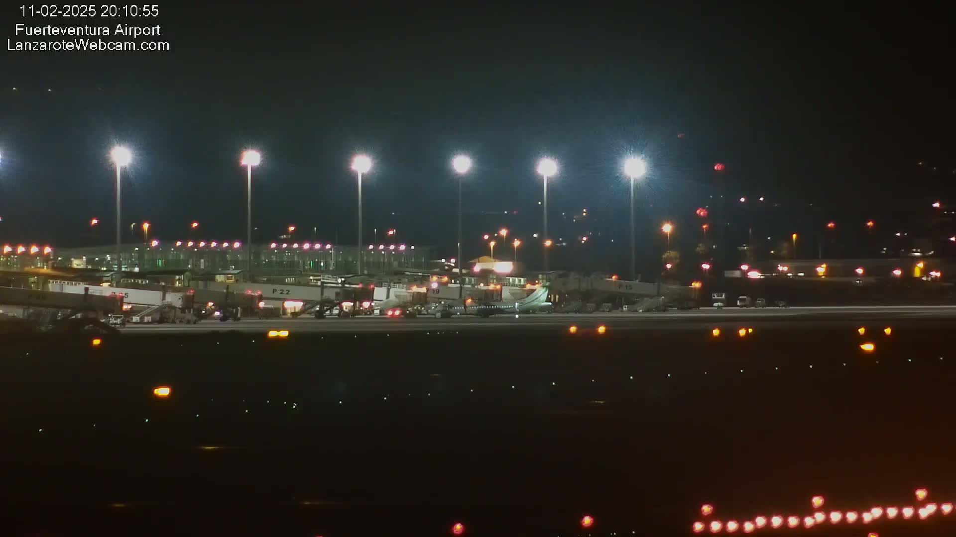 A brightly lit airport at night shows an airplane on the tarmac with ground support vehicles, surrounded by terminal buildings and tall light posts.