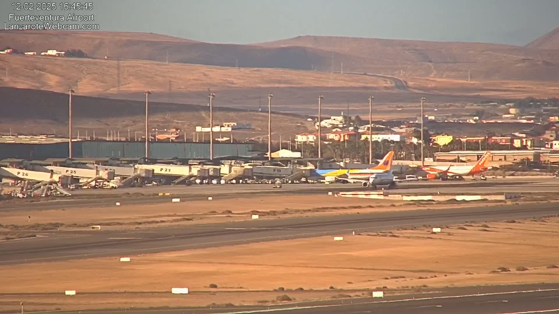 Several airplanes are parked at an airport tarmac under a clear sky, with a dry, hilly landscape visible in the background.