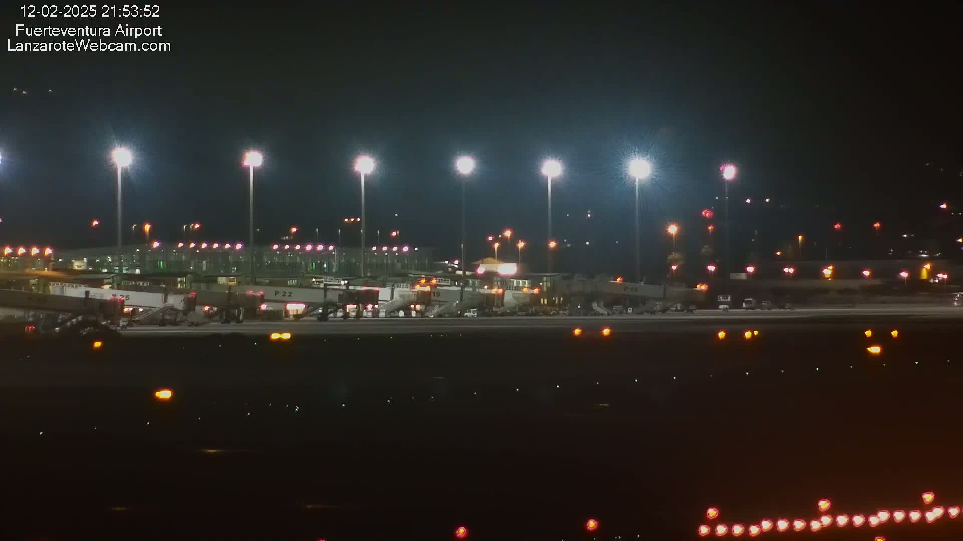 A brightly lit airport terminal at night, with airplanes at the gates and numerous ground vehicles.