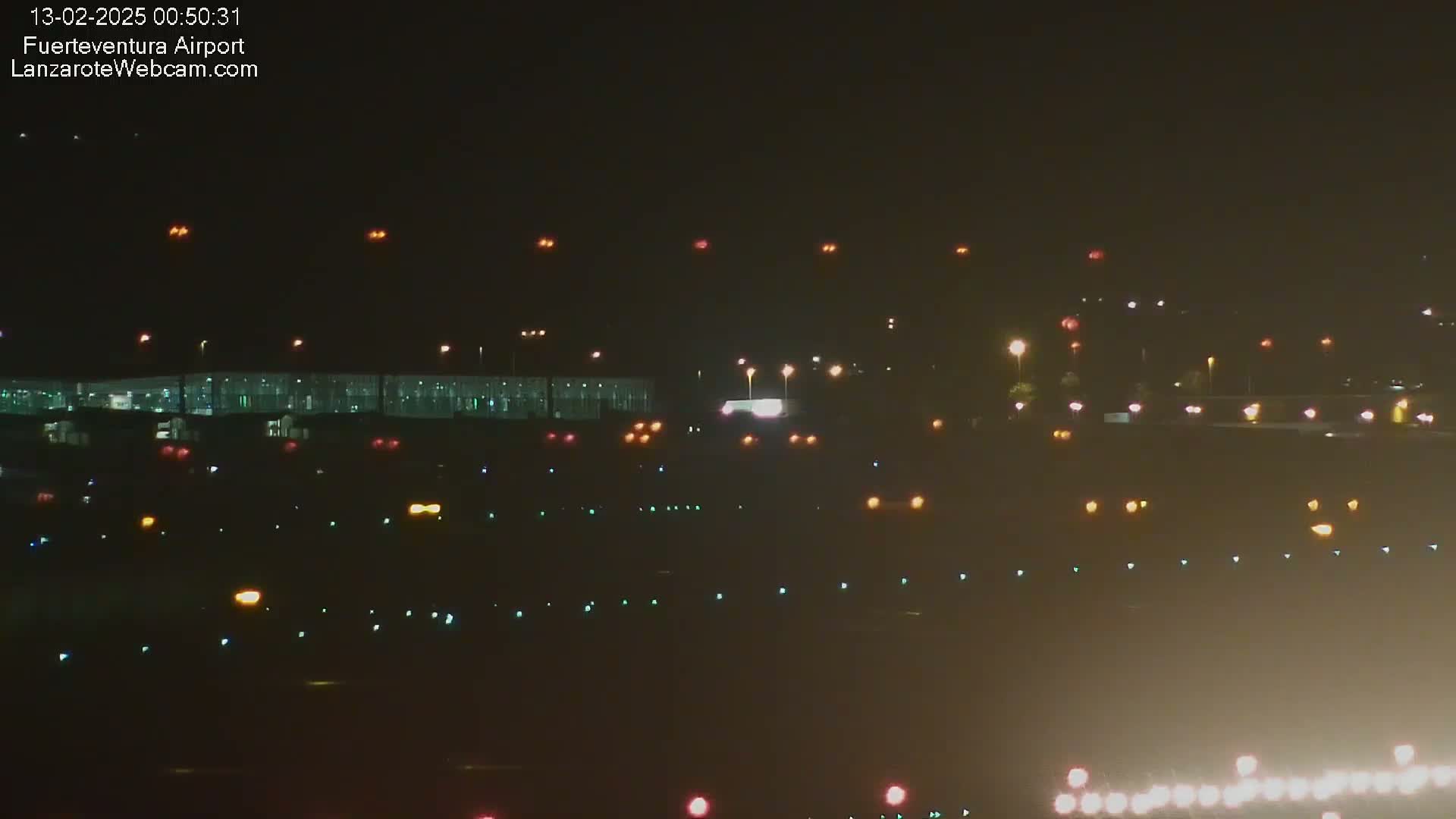 A nighttime long shot of an airport runway and terminal building with numerous lights illuminating the tarmac and surrounding areas under hazy conditions.