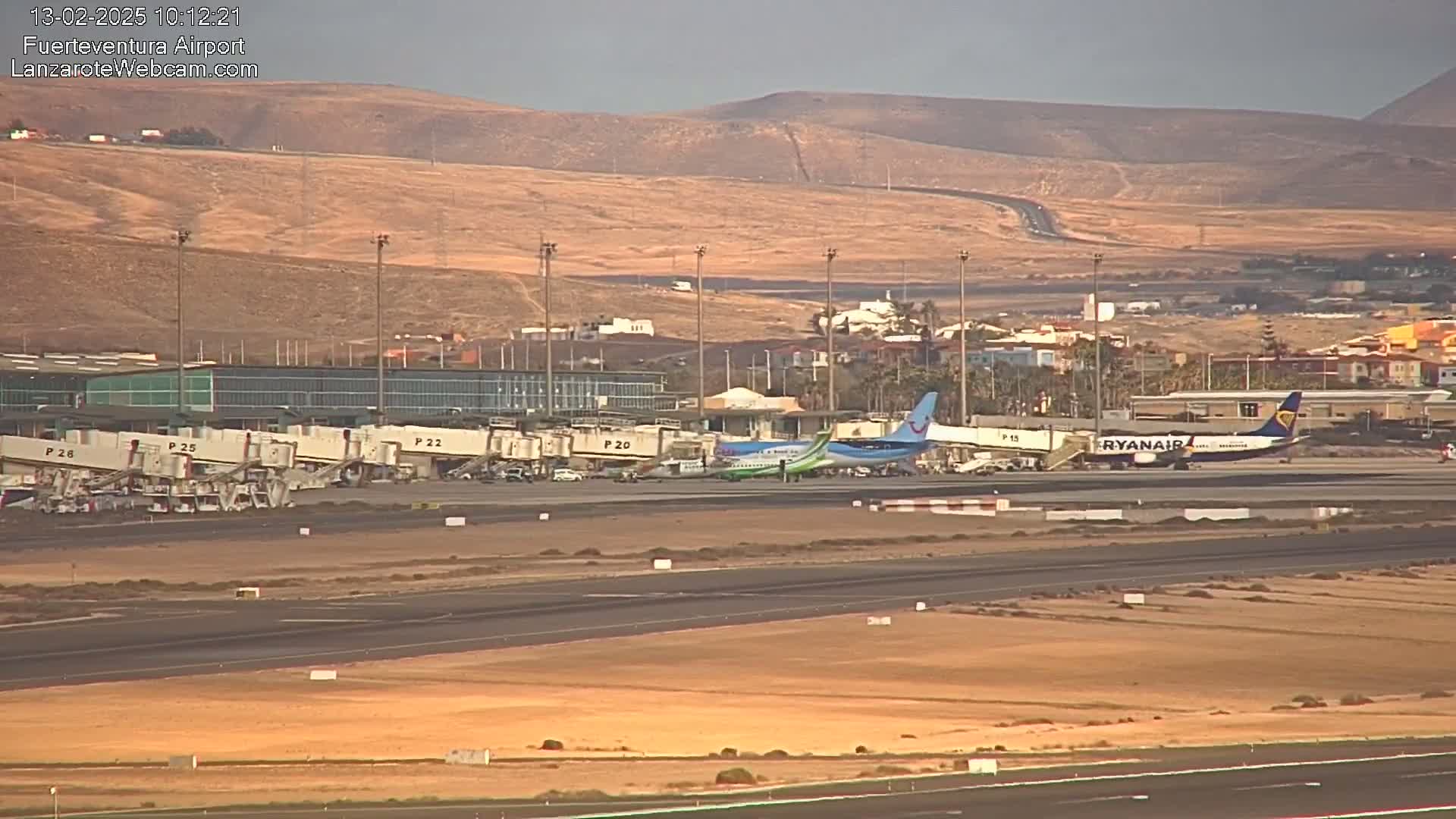 Several airplanes are parked at an airport tarmac near a terminal building, with hills visible in the background under a hazy sky.