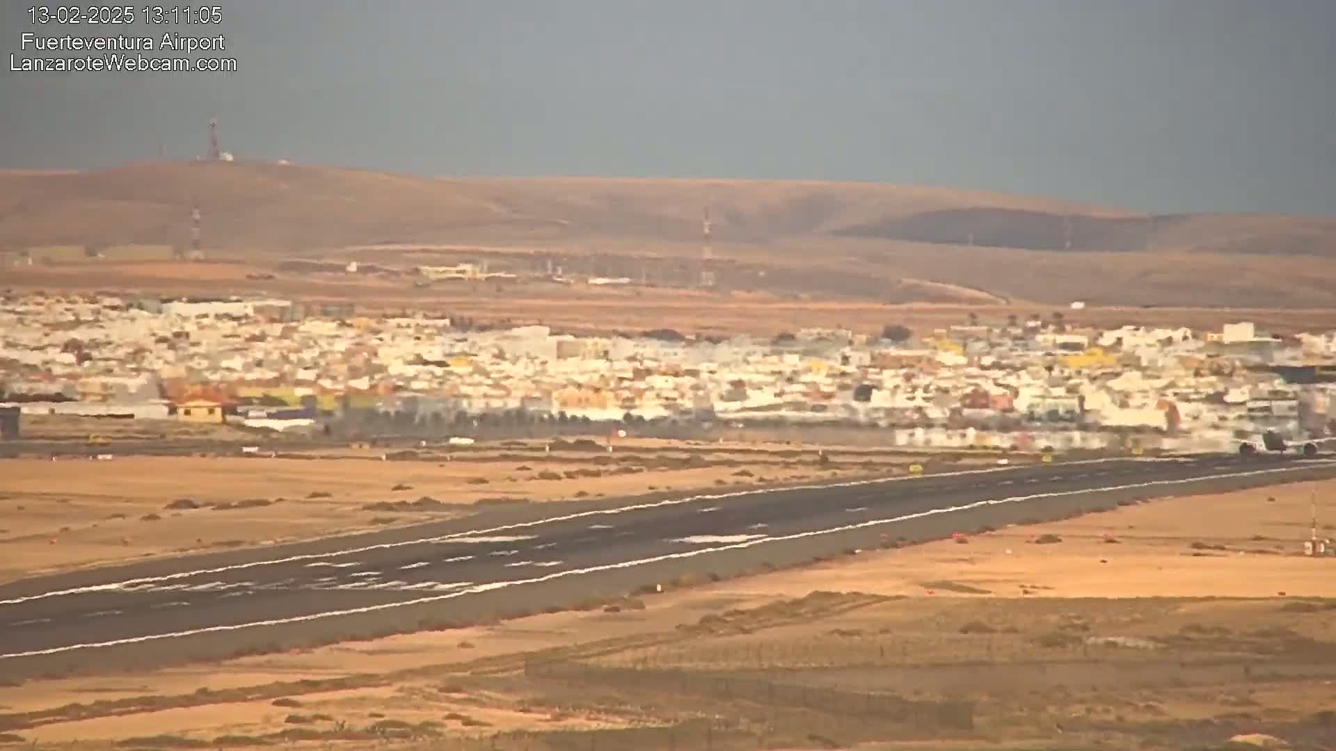An airport runway stretches across a flat, arid landscape toward a distant town under a hazy sky.