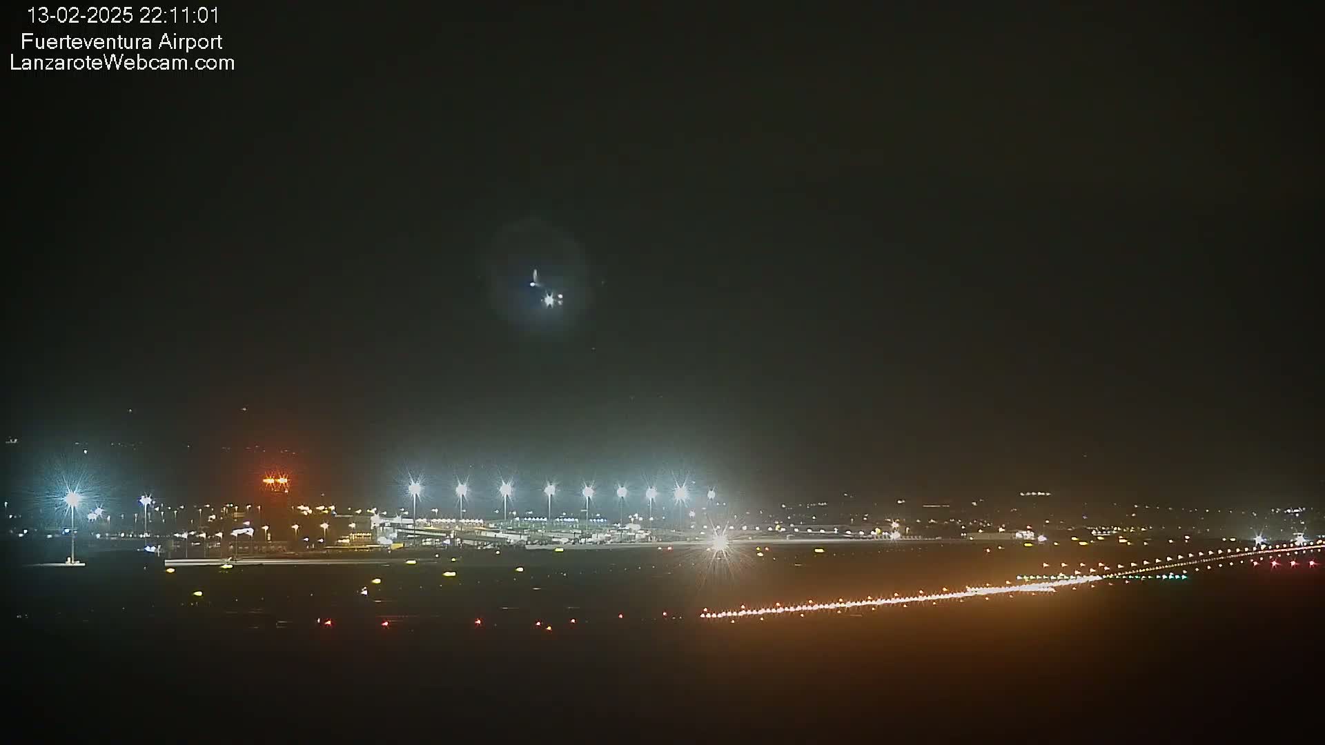 A nighttime long shot of an airport runway with a plane overhead, illuminated by bright lights under a dark sky.