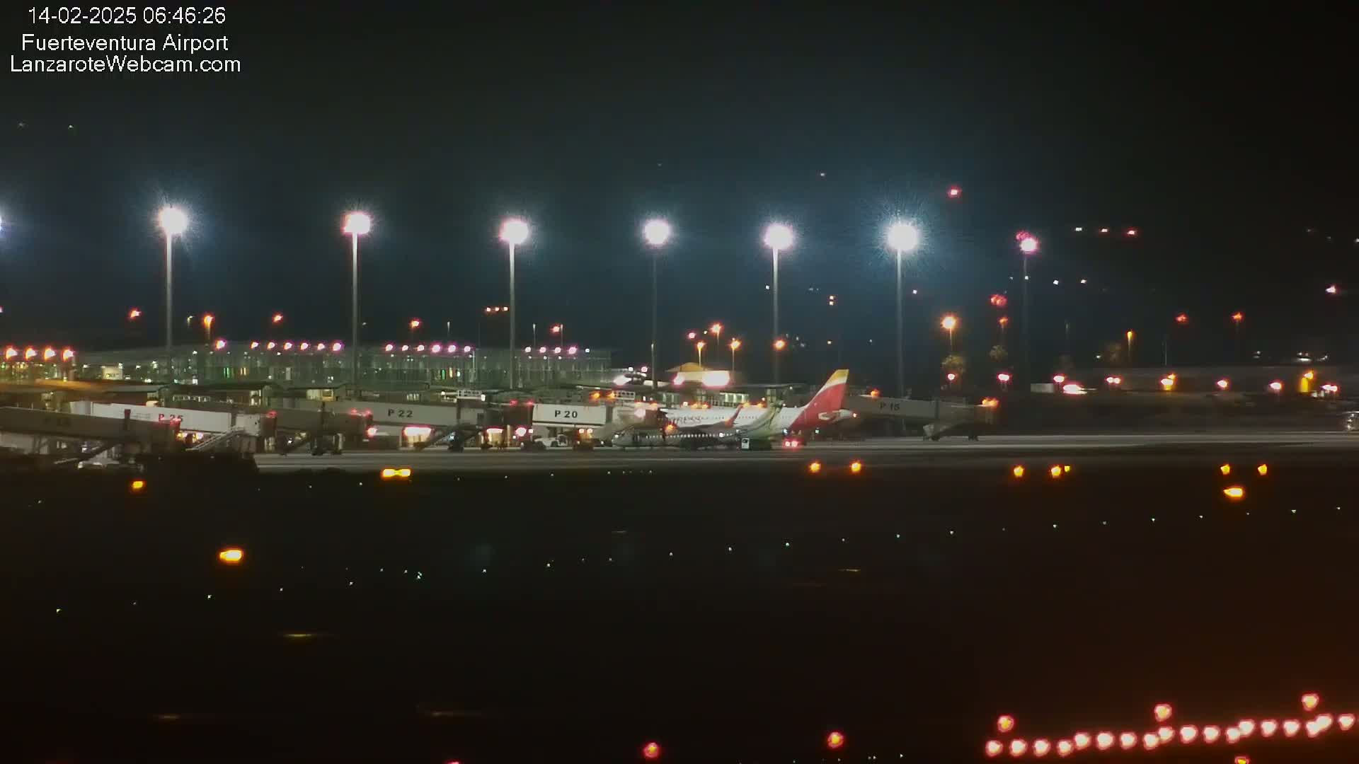 At night, a passenger airplane is parked at a brightly lit airport gate, with other planes and airport buildings visible in the background.