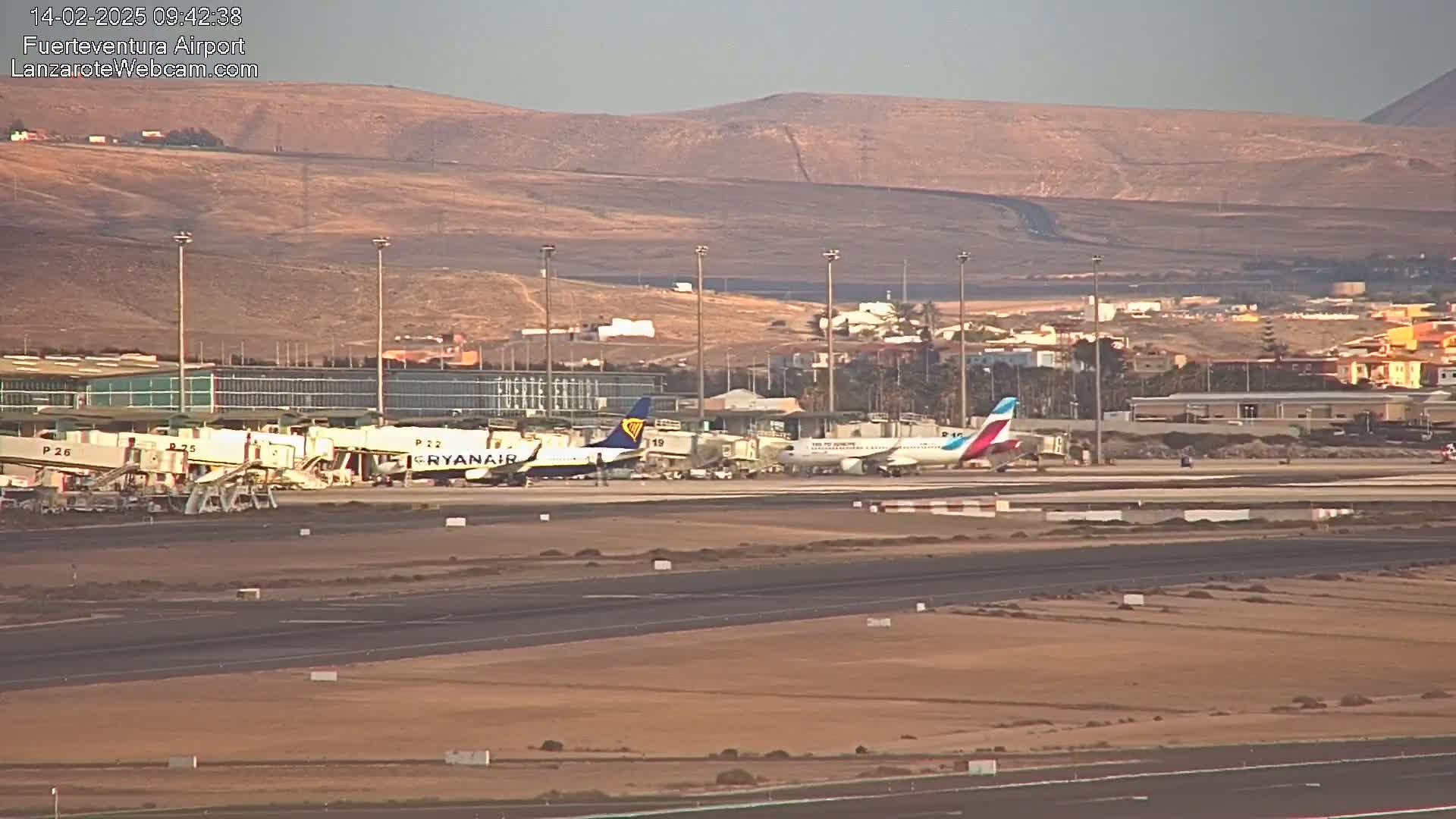 Two airplanes are parked at gates at an airport with a brownish-tan landscape visible in the background under a hazy sky.