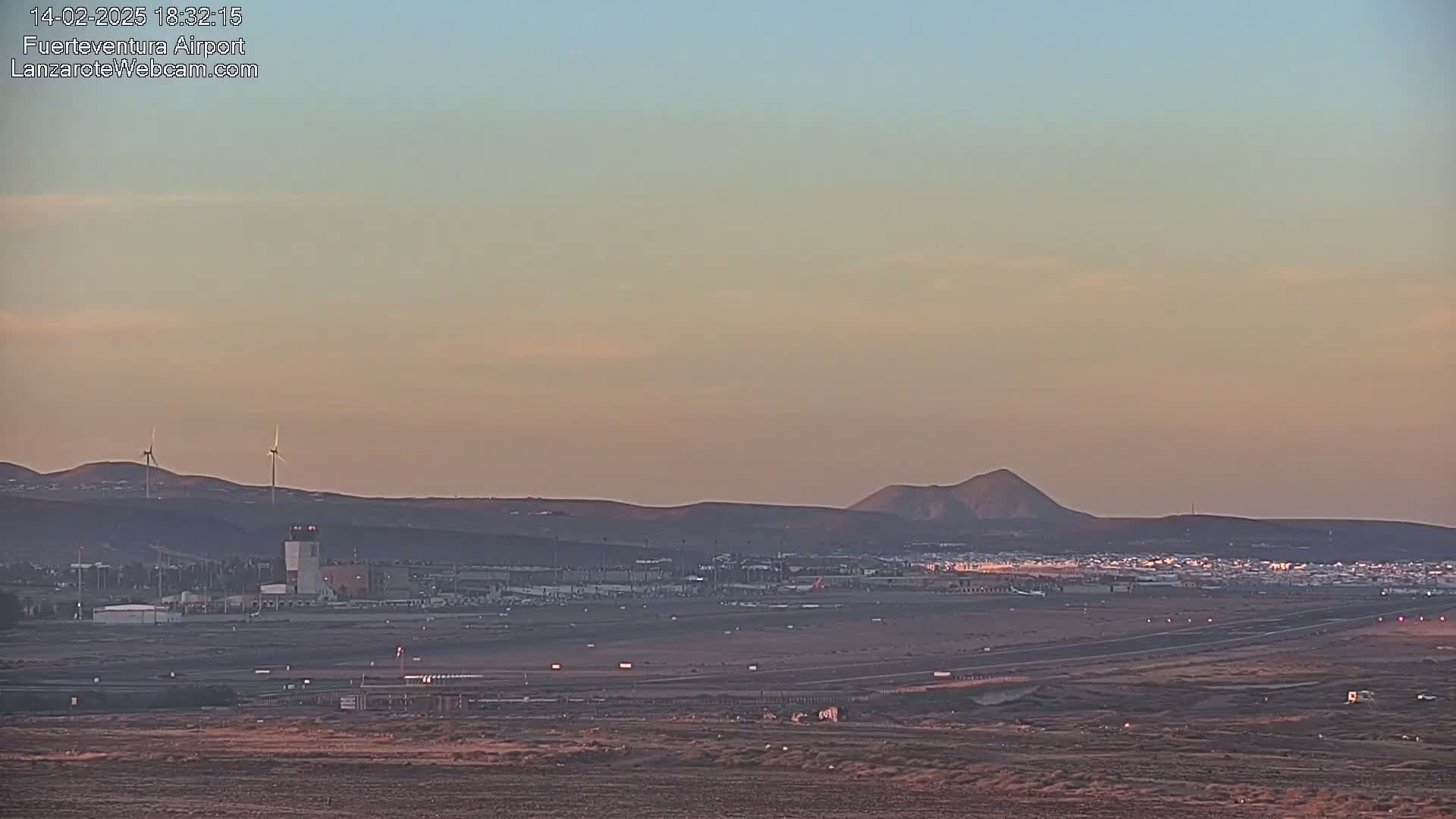 A long shot of an airport at sunset or sunrise under a clear, pale sky, showing a runway, terminal buildings, and wind turbines in the distance.