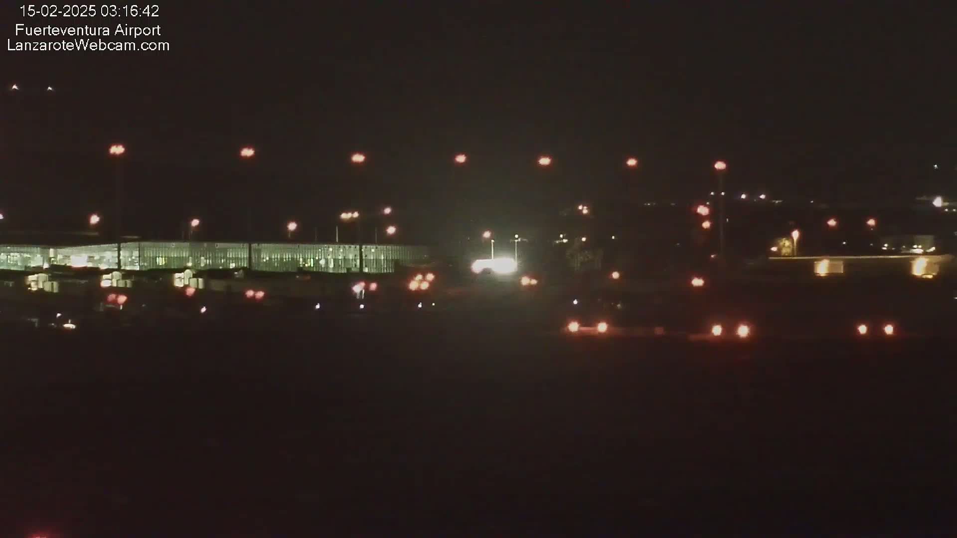 A nighttime long shot of an airport terminal building with various lights illuminating vehicles and the surrounding area under a dark sky.
