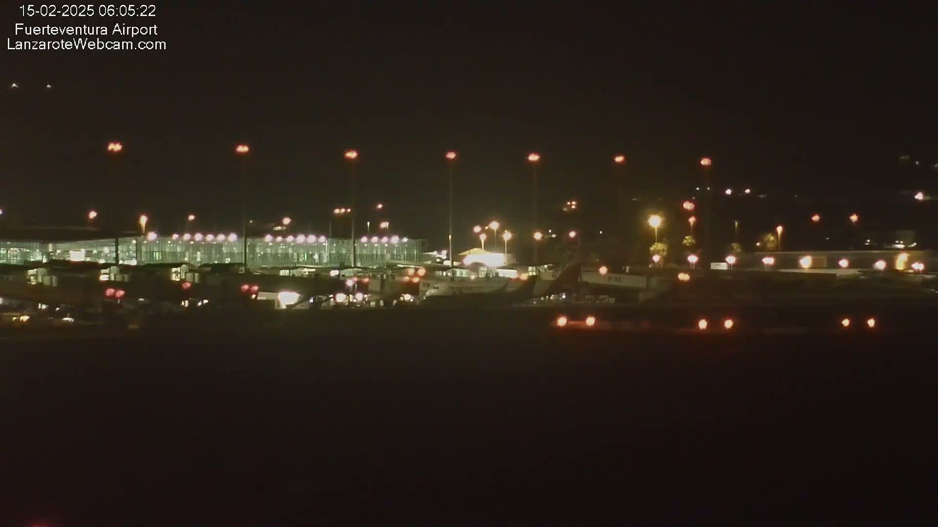 A nighttime view of an airport tarmac with several airplanes parked near brightly lit terminal buildings under a clear, dark sky.