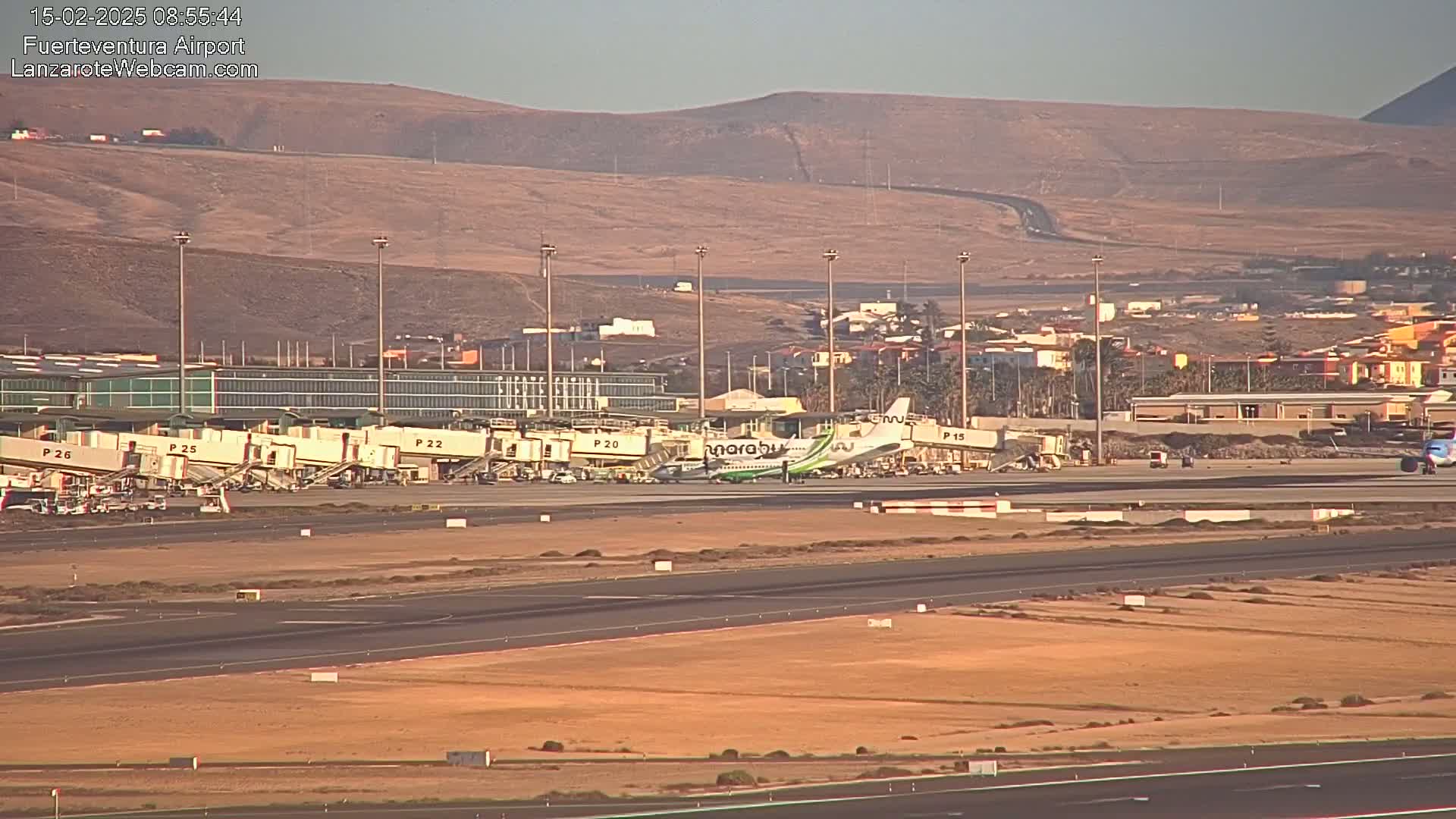 An airplane is parked at a gate at an airport under a clear, sunny sky, with tan-colored hills visible in the background.