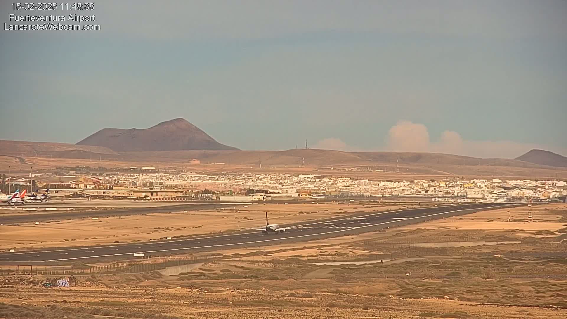 An airplane taxis on a runway at an airport near a town and volcanic hills under hazy, possibly dusty conditions.