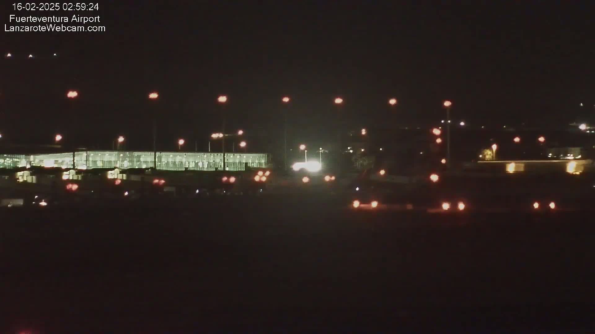 A nighttime long shot of a brightly lit airport terminal building and surrounding roadways with vehicles, under a clear, dark sky.