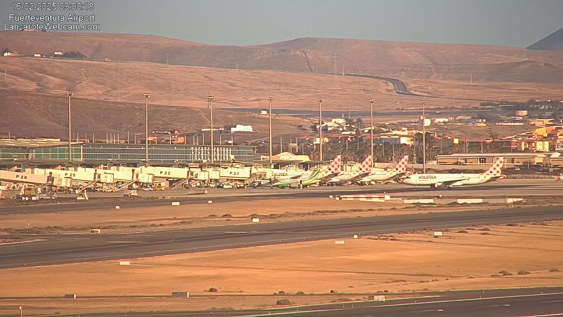 Several airplanes are parked at gates at an airport under a clear, sunny sky, with dry, brown hills visible in the background.