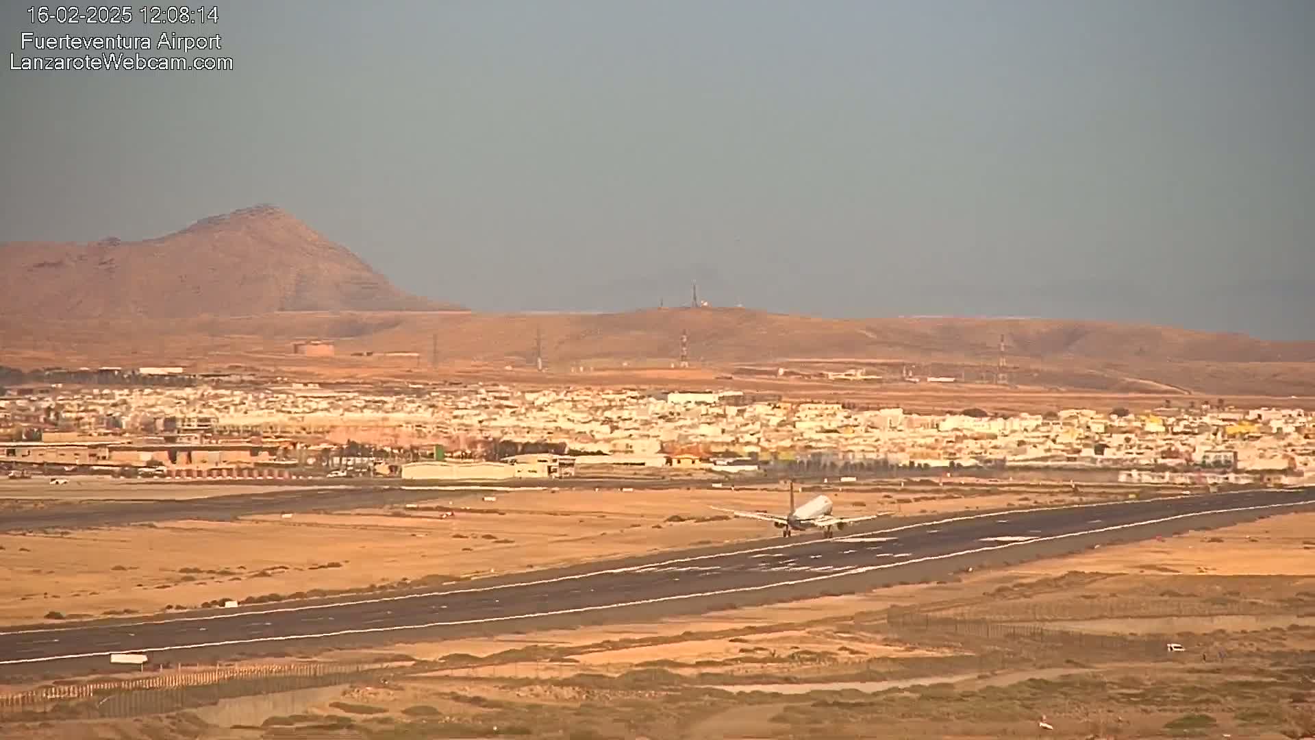 A passenger plane lands on a runway near a town situated in a desert-like landscape under a hazy sky.