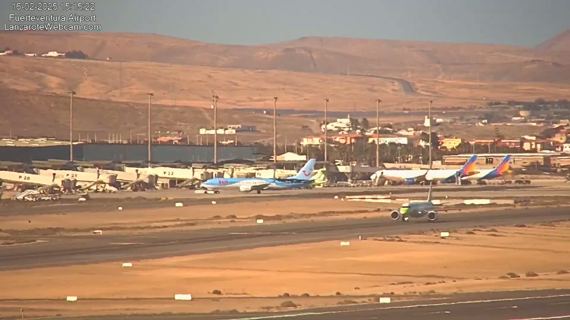 Several airplanes are parked and taxiing on a tarmac at an airport under a hazy, sunny sky, with a dry, hilly landscape visible in the background.