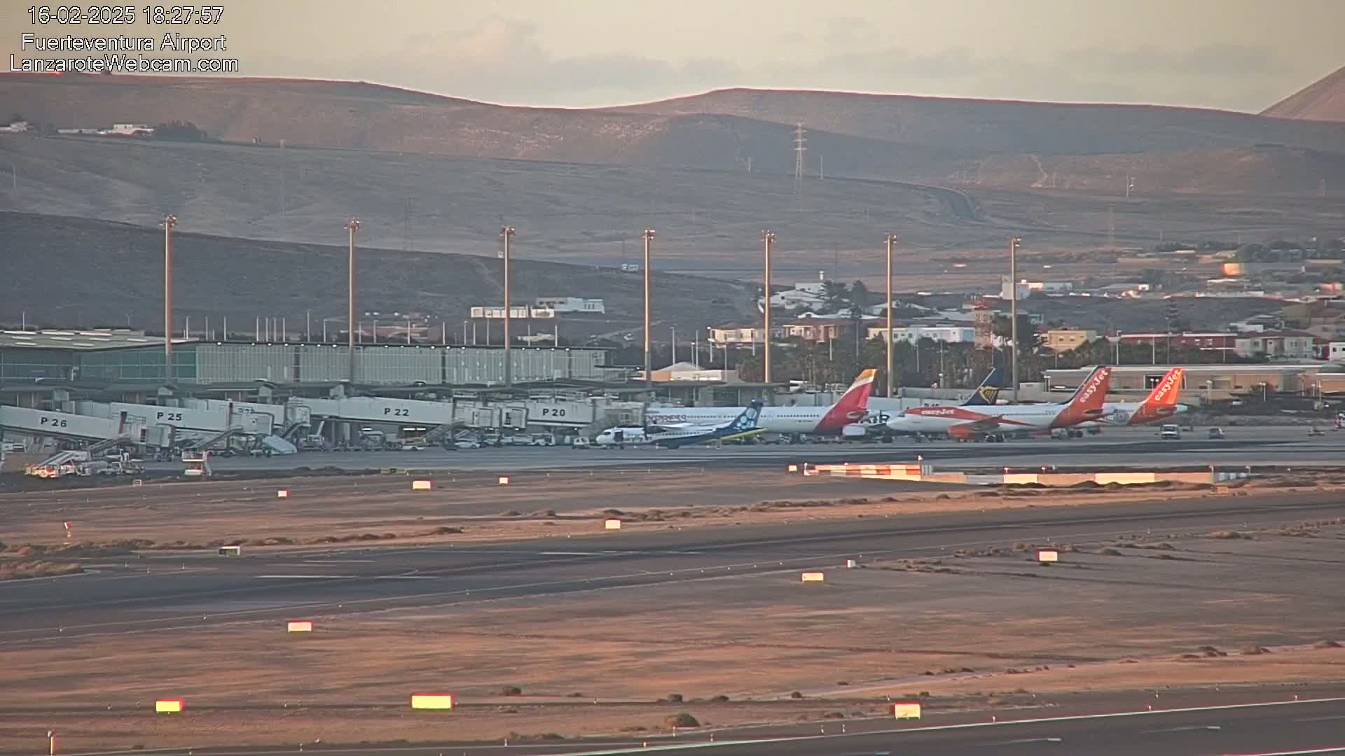 Several airplanes are parked at an airport tarmac near a terminal building under a clear, dusky sky with distant hills in the background.