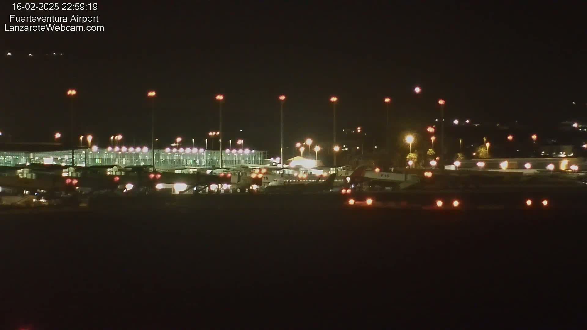 At night, an airport terminal and tarmac are illuminated, showing several aircraft and ground vehicles.