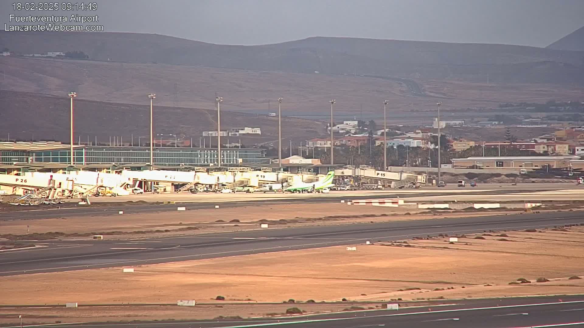 An airport tarmac with several airplanes at gates, surrounded by airport buildings and a hazy, distant mountain range.