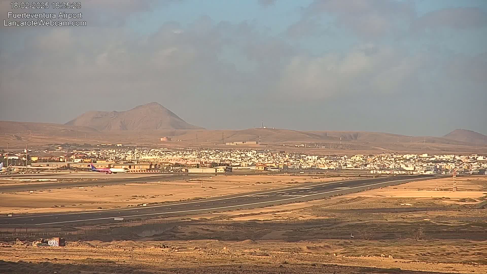 An airplane taxis on a runway at an airport, with a town and desert landscape under a partly cloudy sky.