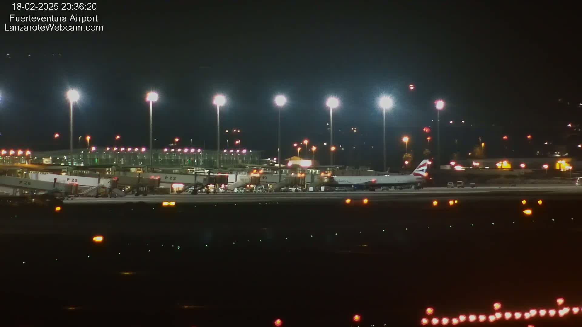 A brightly lit airport at night shows an airplane parked at a gate, with several other gates visible in the background.