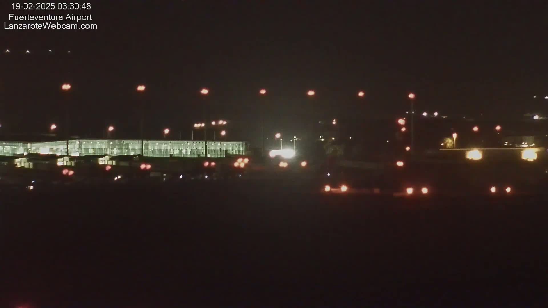 A brightly lit airport terminal building at night is visible, with various vehicles' lights scattered in the foreground and background.