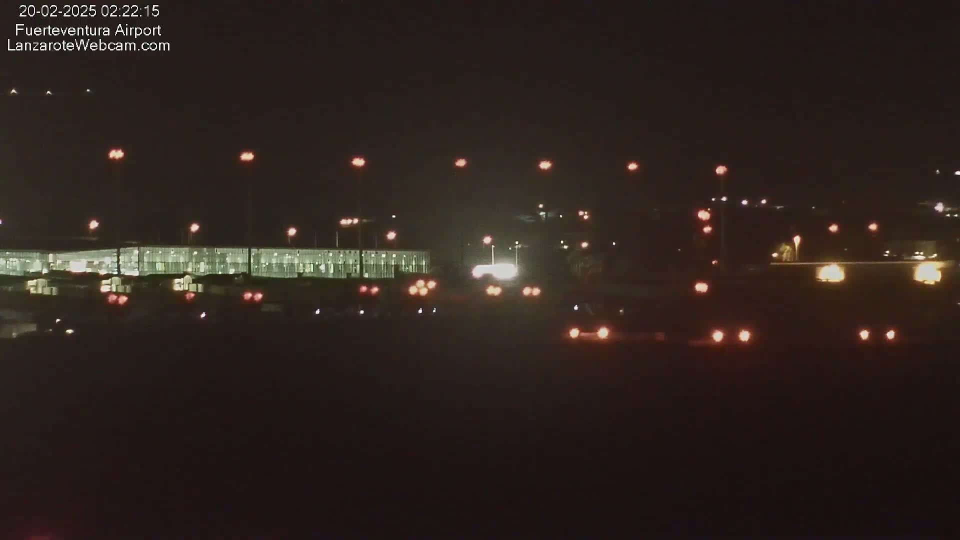 A brightly lit airport terminal is visible at night, with numerous lights illuminating vehicles and the surrounding area.