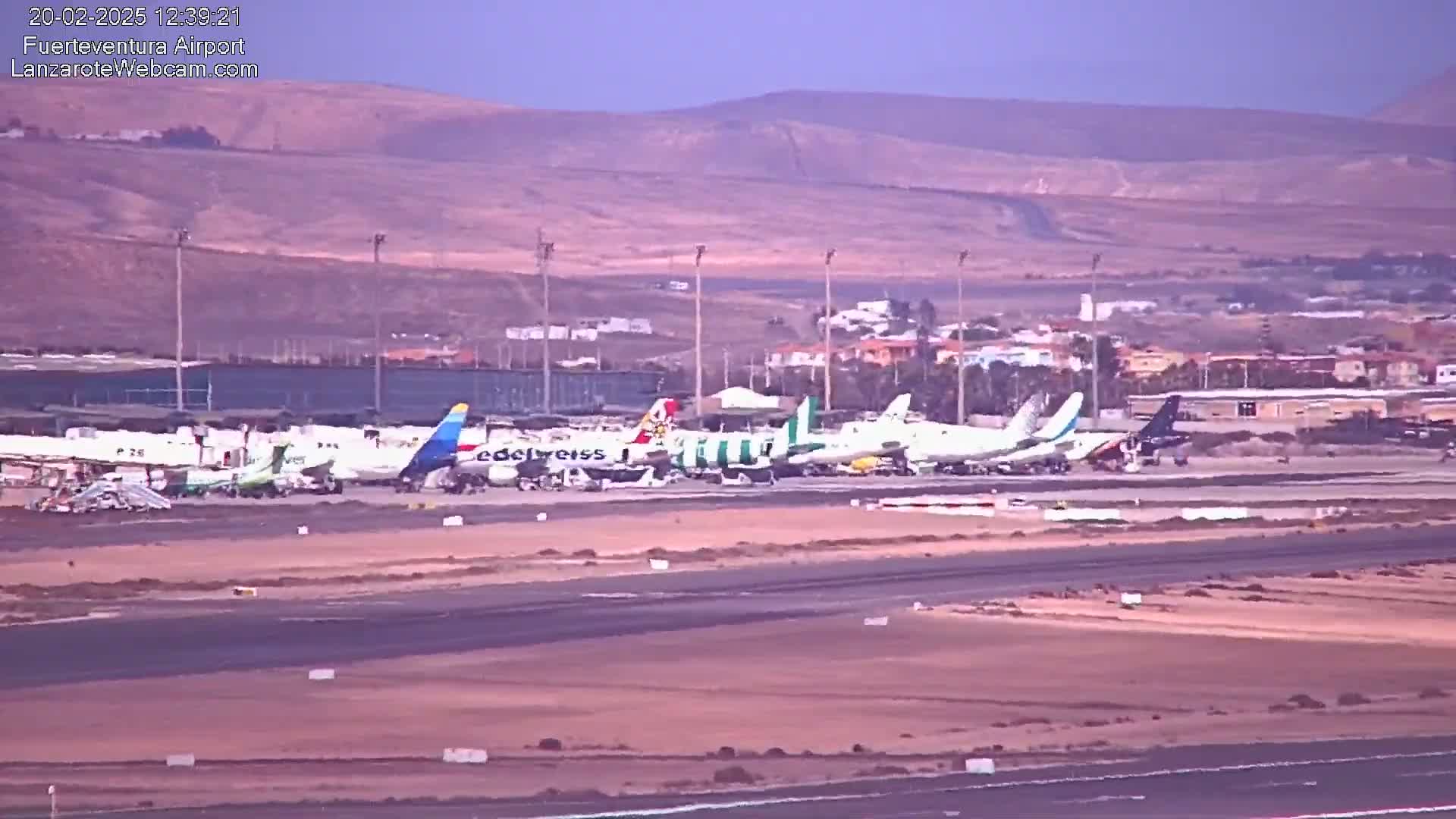 Several airplanes are parked on an airport tarmac under a hazy sky, with brown hills visible in the distance.