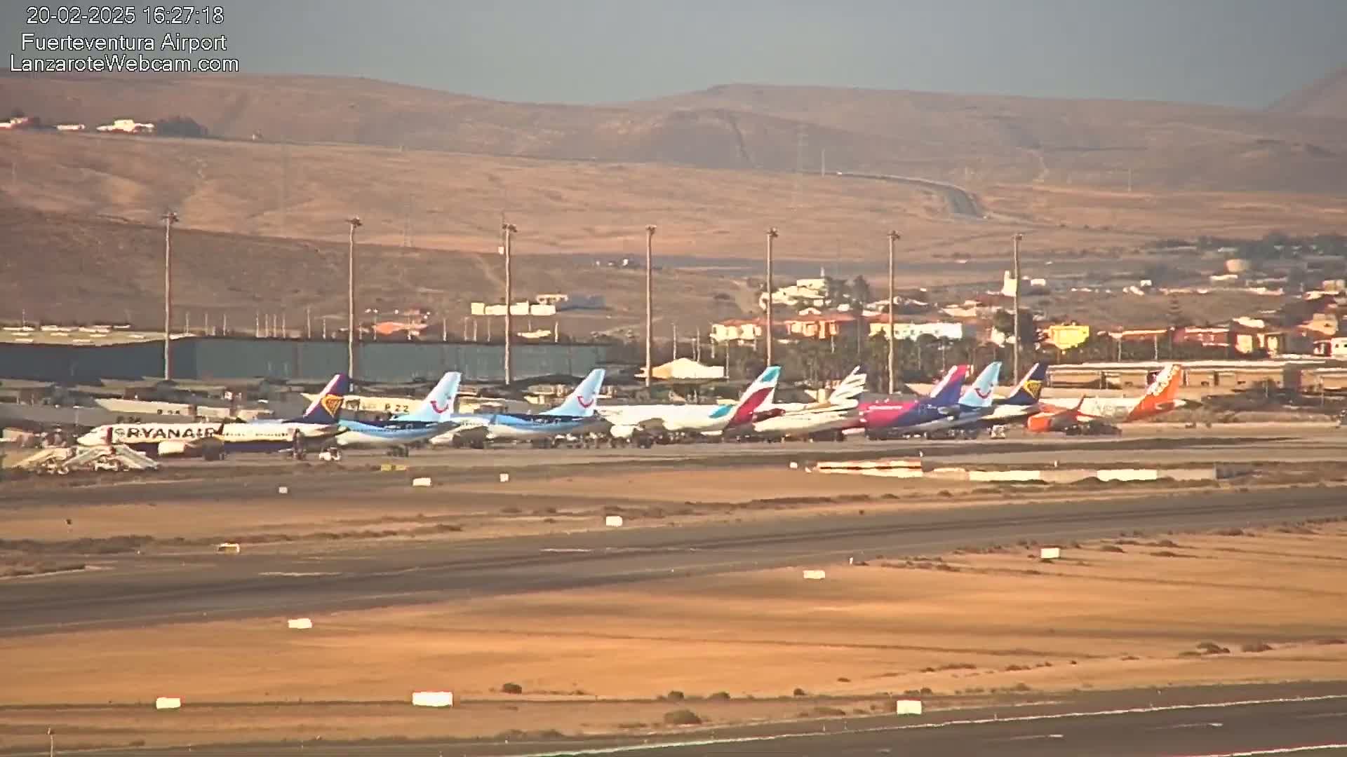 A row of various passenger airplanes are parked on a tarmac at an airport under hazy, sunny conditions, with dry, brown hills visible in the background.