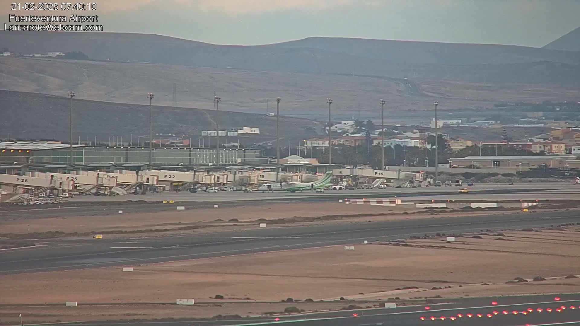 A plane is parked at a terminal in an airport with several jet bridges, under an overcast sky, with mountains in the background.