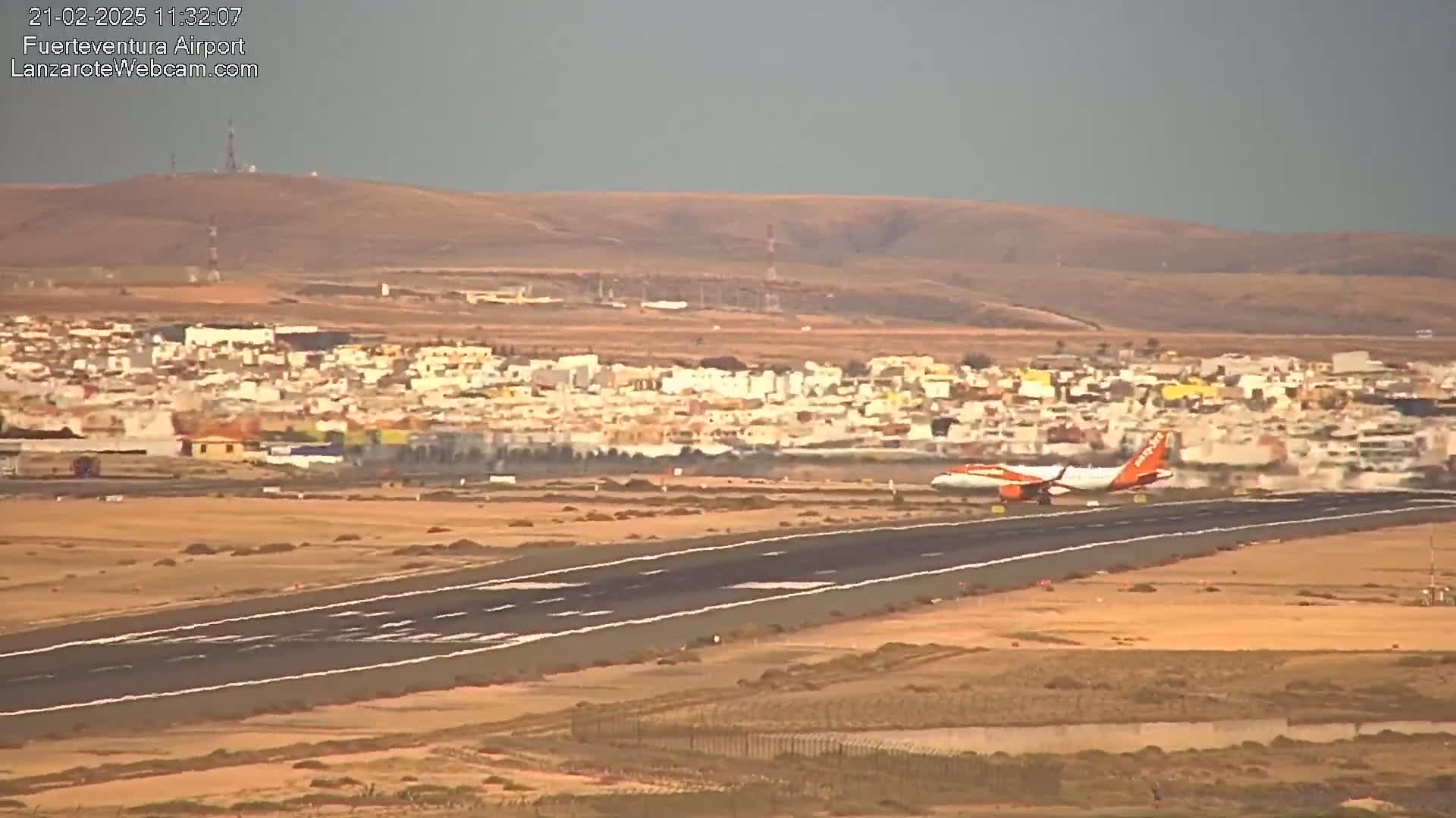 An orange and white airplane taxis on a runway near a town under hazy, sunny conditions.