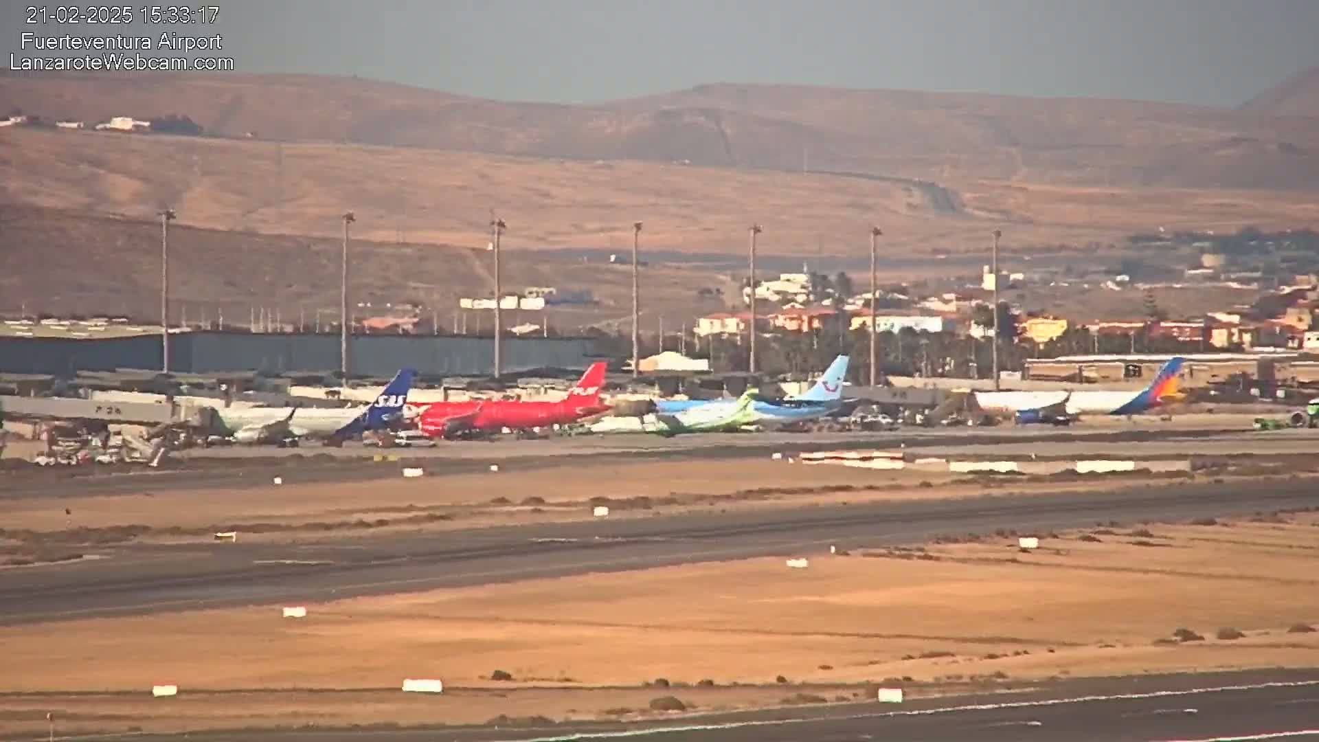 Several airplanes are parked on a tarmac at an airport, with a hilly, desert-like landscape visible in the background under a clear sky.