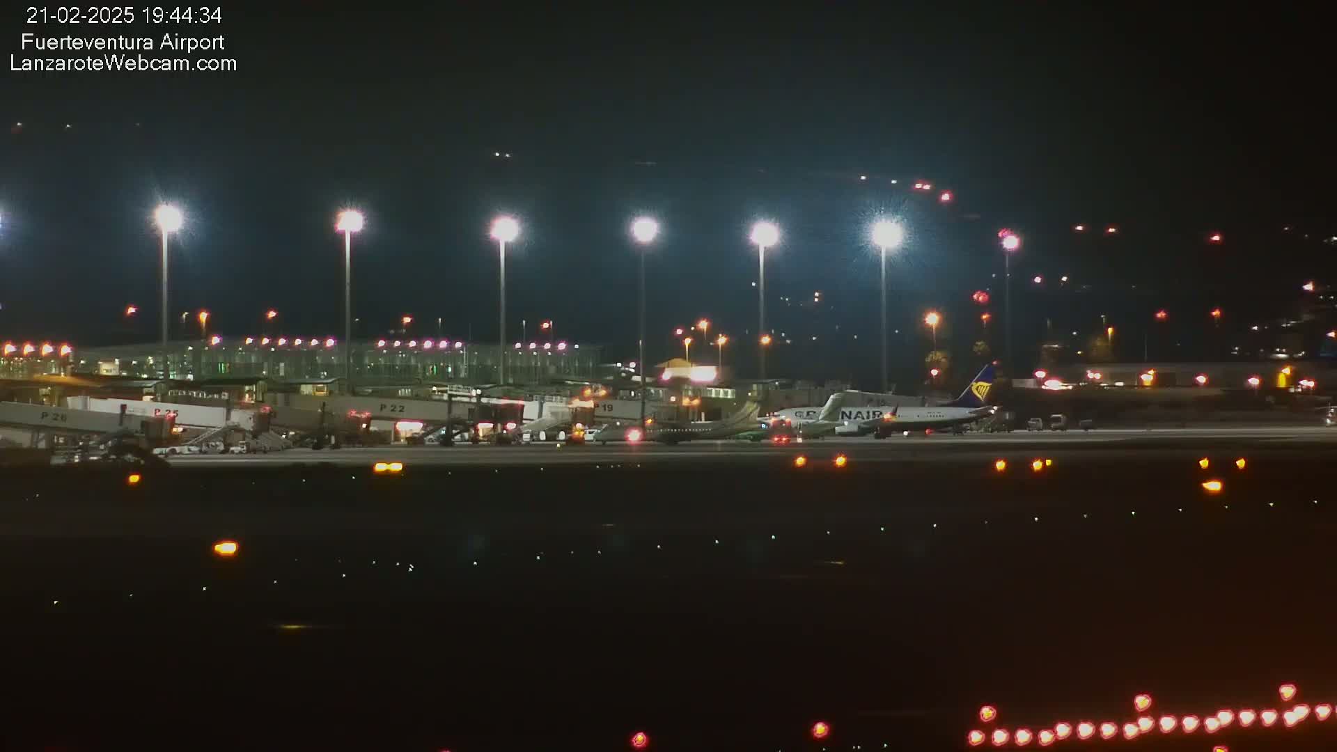 At night, a brightly lit airport tarmac shows several airplanes near gates and passenger boarding bridges, under floodlights.